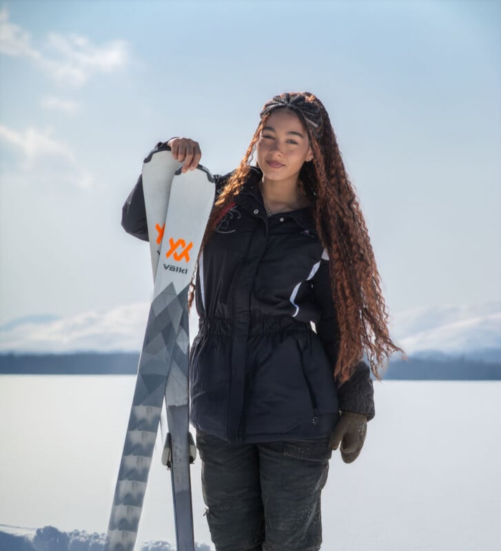A woman with long curly hair stands in the snow, smiling and holding a pair of skis upright. She wears a black winter jacket and gloves, with snowy mountains and a bright sky in the background.