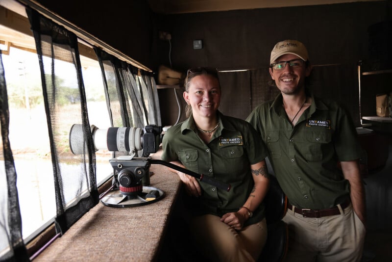 Two people in safari uniforms with "Untamed" logos smile inside a safari vehicle. One sits next to a professional camera on a tripod pointing out the window; the other stands beside them. Light streams in from large windows.