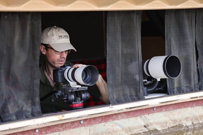 A man wearing glasses and a cap uses a camera with a large telephoto lens, positioned at a window with black curtains. Another camera with a similar lens rests nearby.