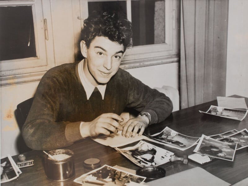 A young man smiles while sitting at a desk covered with black-and-white photographs and papers, holding a pen. He appears to be working or writing, with windows and curtains in the background.