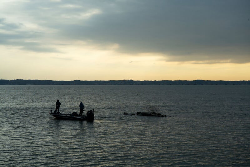 A small boat with two people fishing on a calm lake during sunset, with a cloudy sky and distant shoreline visible in the background.