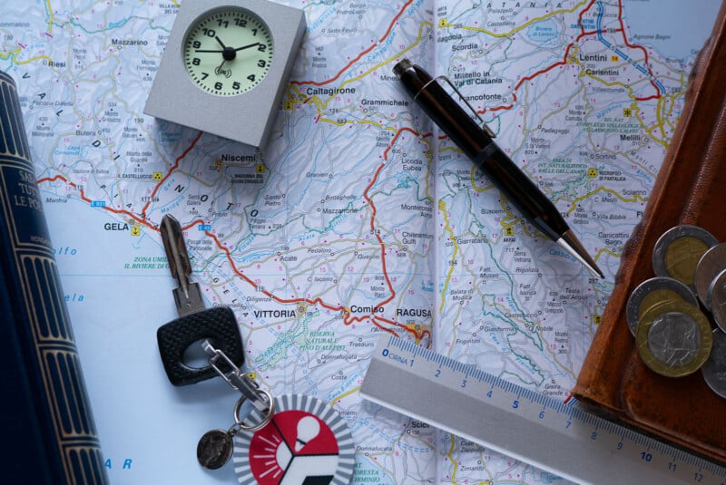 A map of Sicily with a pen, a ruler, a clock, euro coins in a wallet, a set of keys, and a book placed on top, suggesting planning a trip or journey.