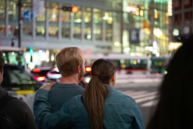 A couple stands close together, facing a busy city street at night with bright lights and blurred cars passing by. The woman has her arm around the man's shoulder, and both wear denim jackets.