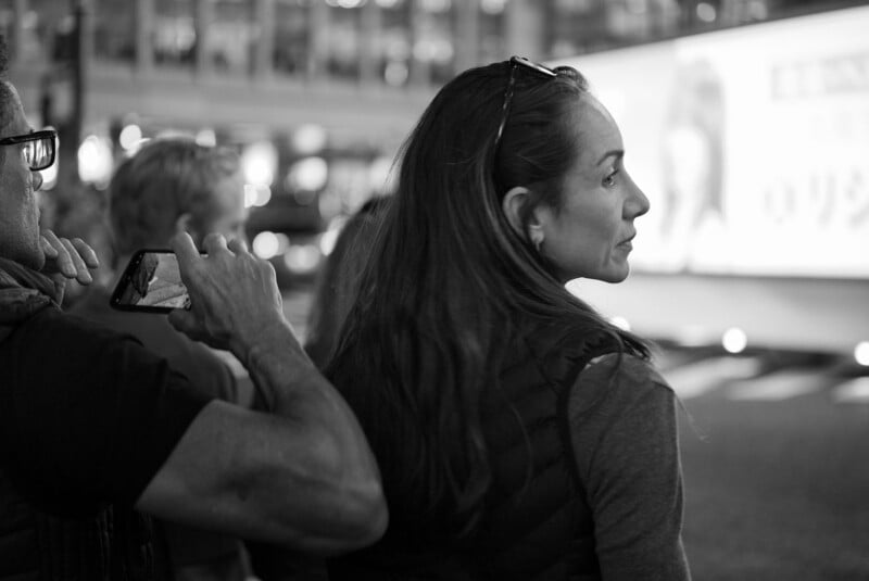 A woman with long hair and sunglasses on her head looks to the side in an urban setting at night. A man beside her holds up a phone, taking a photo. The scene is in black and white, with blurred lights in the background.