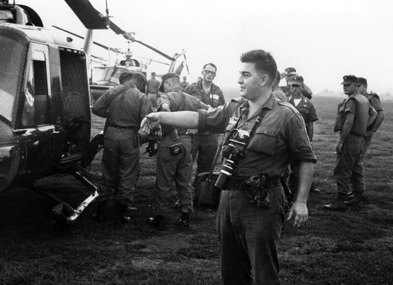 A man with cameras around his neck gestures while standing beside a helicopter. Several soldiers in uniform stand nearby, some boarding the helicopter. The scene appears to be from a military setting.