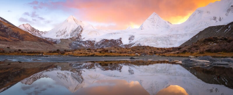 Snow-capped mountains are reflected in a calm lake at sunset, with orange and pink clouds lighting the sky above rocky terrain and grassy land in the foreground.