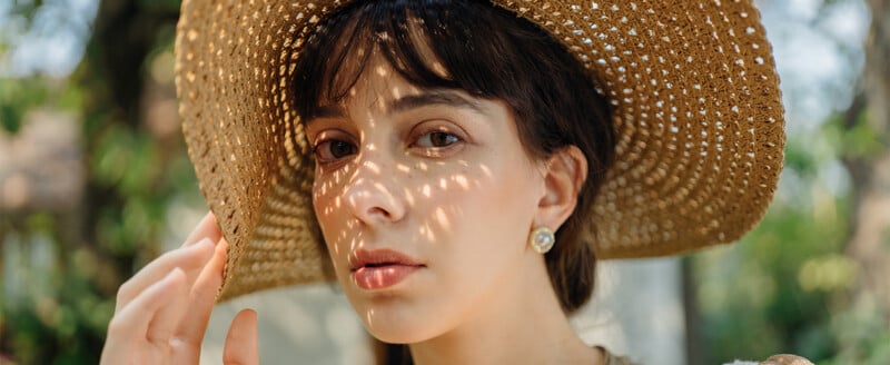 A woman wearing a wide-brimmed straw hat stands outdoors, with sunlight creating dappled patterns on her face. She has long dark hair, wears earrings, and looks directly at the camera with a calm expression.