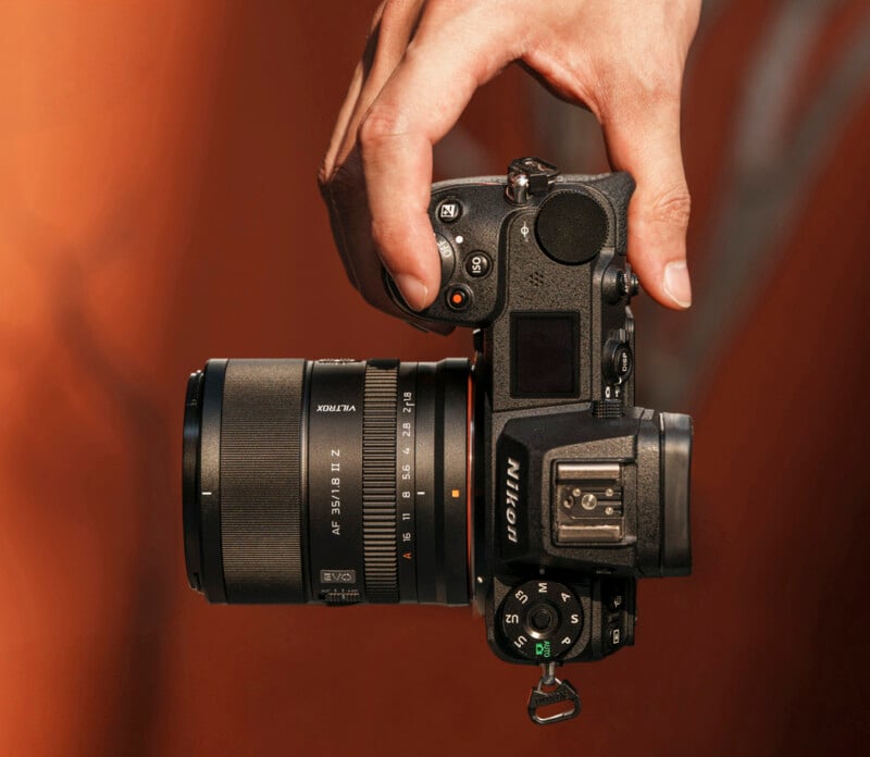 A person’s hand holds a black Nikon mirrorless camera with a large lens, set against a blurred, warm brown background.