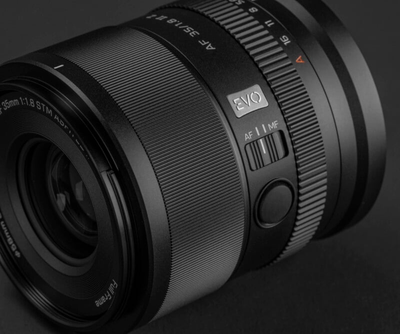 Close-up of a black camera lens showing detailed texture, focus ring, AF/MF switch, and "EVO" label, all set against a dark background.