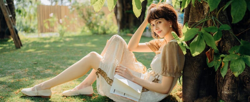 A young woman in a light dress sits on grass, leaning against a tree in a sunlit garden, holding an open book and looking at the camera. Sunlight filters through the leaves, creating a peaceful atmosphere.