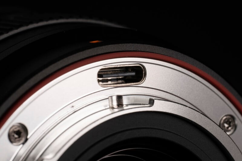 Close-up of a camera lens mount with a metallic ring, screws, and electronic contacts, showing detailed texture and a small orange alignment mark against a black background.