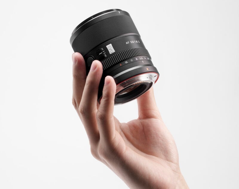 A hand holding a black camera lens with visible focus and aperture markings, against a plain white background.