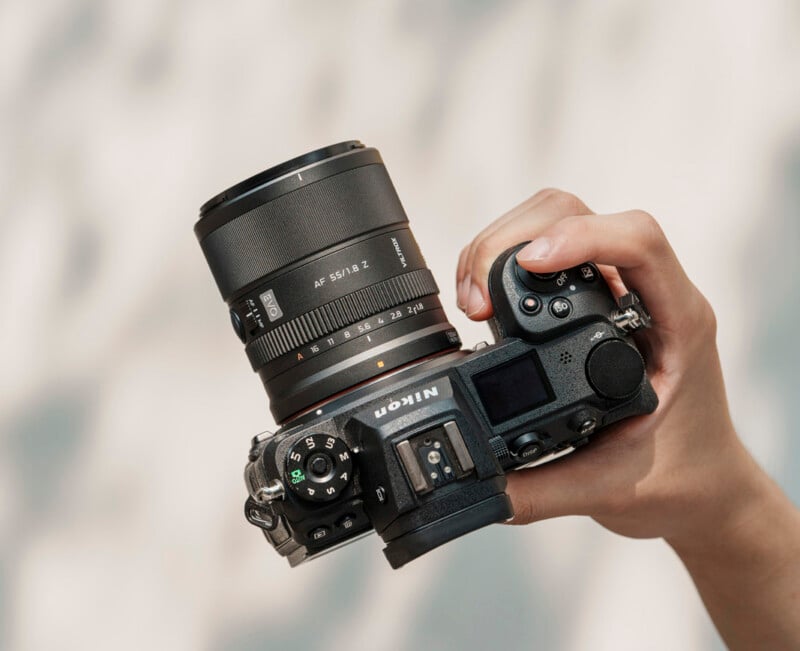 A hand holding a Nikon digital camera with a 50mm lens against a blurred, light-colored background. The camera is viewed from above, showing its buttons and dials.