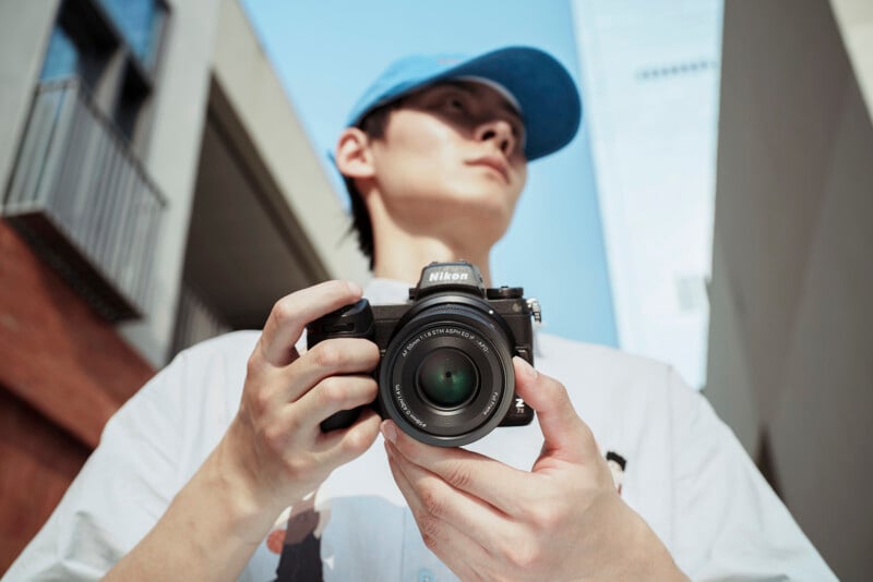 A person wearing a blue cap and white shirt holds a Nikon camera, looking upward, with urban buildings and a blue sky in the background. The photo is taken from a low angle.