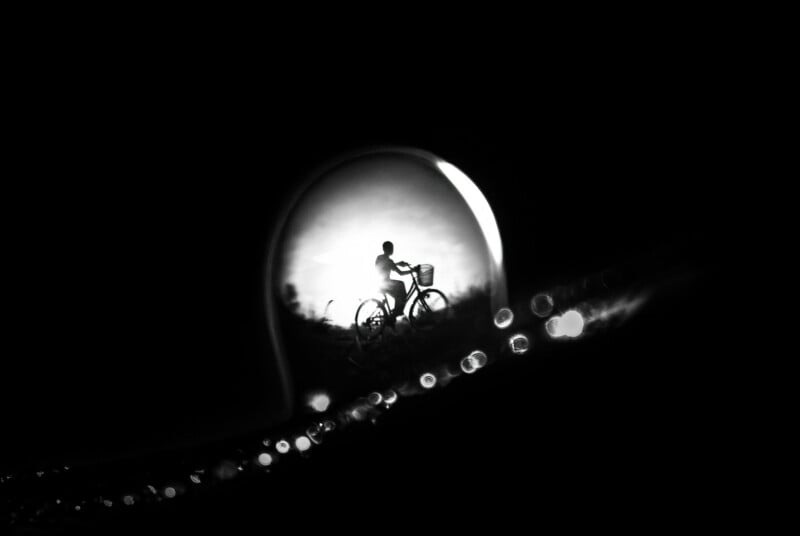 A silhouette of a person riding a bicycle, framed by a lightbulb shape in darkness, with glowing bokeh dots along the lower edge, creating a dreamy, surreal atmosphere.