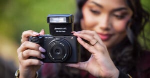 A woman with manicured nails holds a Sony camera with a Westcott 518 Mini Flash attached, focusing on the camera while the background is softly blurred.