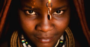 Close-up of a person with braided hair and beaded necklaces, looking directly at the camera. A decorative chain hangs down the center of their forehead, and warm lighting highlights their features.