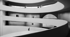 Black and white photo of a modern, spiral interior with curved ramps and walls. Three people walk on different levels, emphasizing the architectural lines and light streaming in from windows above.