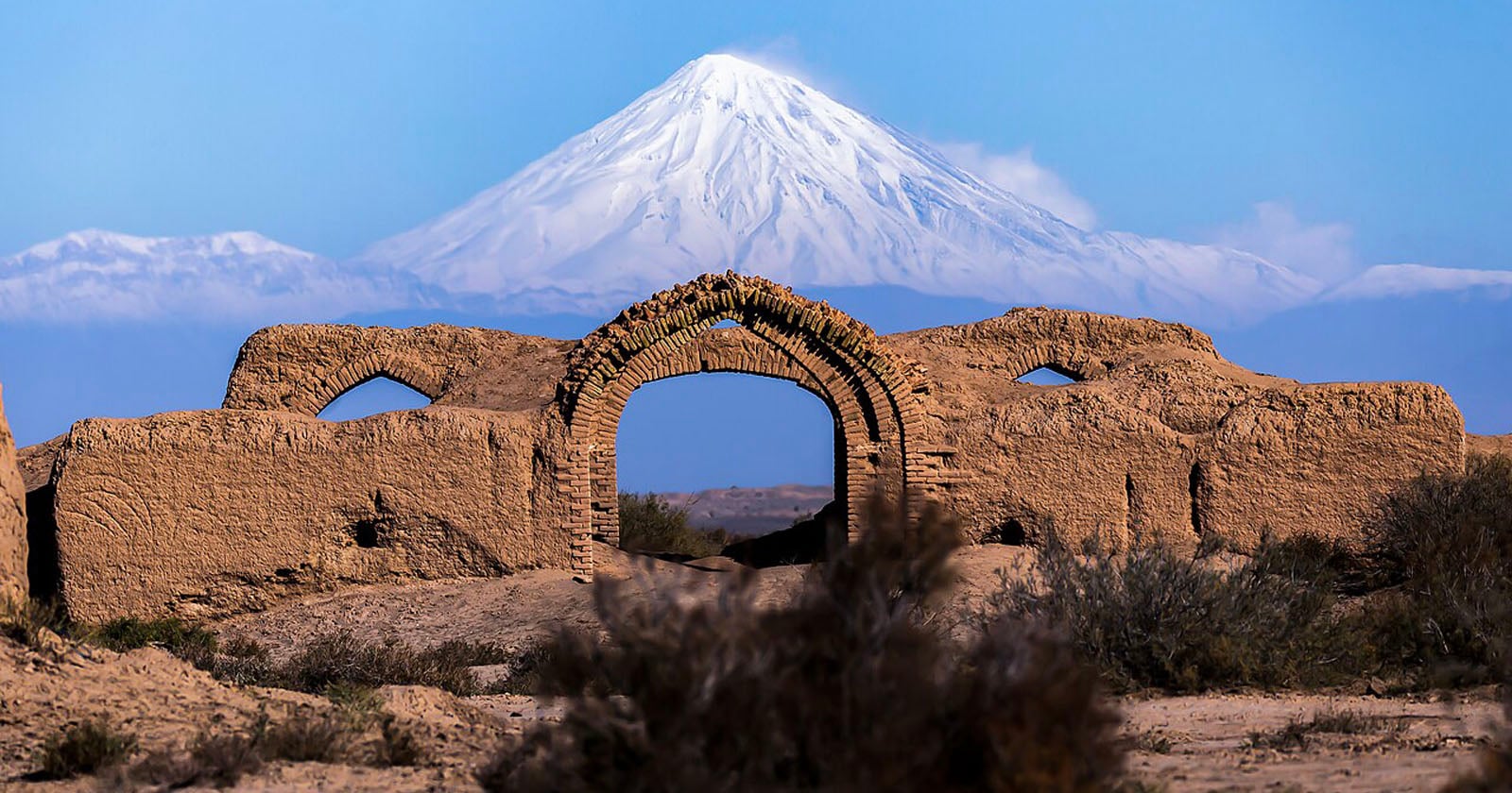 An ancient, sandy brick archway stands in the foreground, with crumbling walls on each side. In the background, a tall snow-capped mountain rises majestically against a clear blue sky.