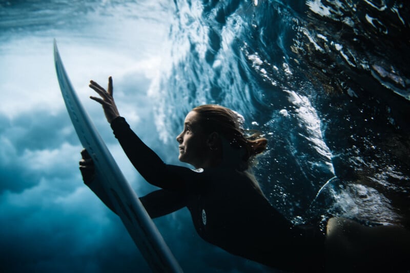 A surfer underwater in a wetsuit holds onto a surfboard, swimming beneath a wave with sunlight filtering through the water above. The scene captures movement and intensity beneath the ocean surface.