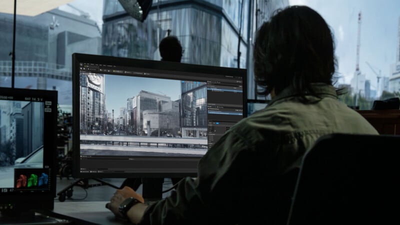 A person sits at a desk, working on a computer with a large monitor displaying a cityscape image in editing software. Reflections of modern buildings can be seen through large windows behind them.