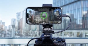 A smartphone mounted on a camera is displaying a cityscape with a digital overlay of hexagonal guides. Both devices are set up on a balcony with tall glass buildings in the background.