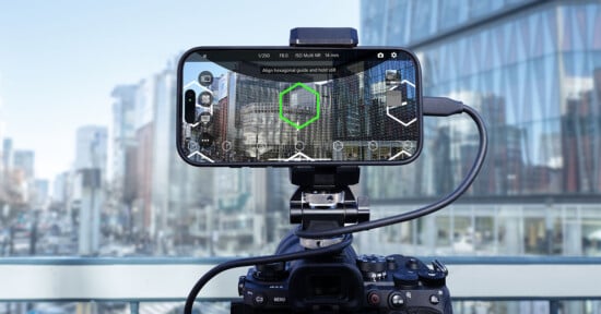 A smartphone mounted on a camera is displaying a cityscape with a digital overlay of hexagonal guides. Both devices are set up on a balcony with tall glass buildings in the background.