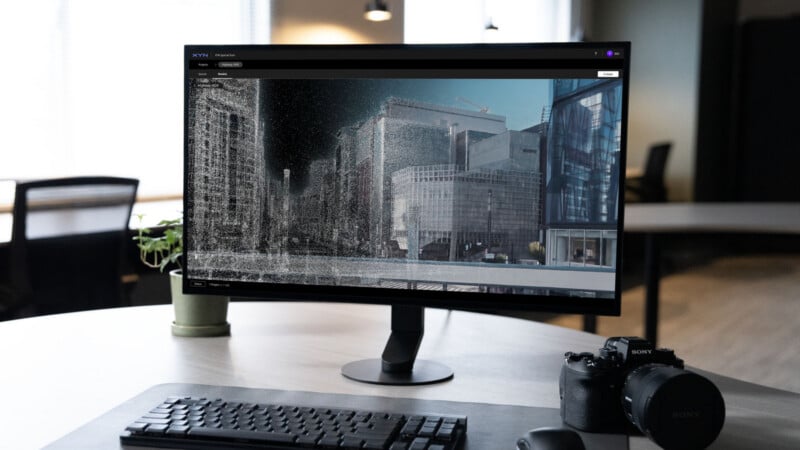 A computer monitor on a desk displays city buildings rendered in 3D, with a keyboard, mouse, Sony camera, and potted plant nearby in a modern, softly lit office setting.