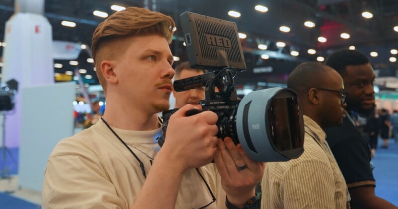 A person holding a professional video camera films at an indoor event, with blurred people and bright lights visible in the background.