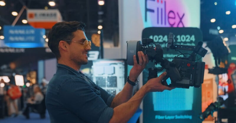 A man in sunglasses holds and films with a large video camera at a tech or media expo, with colorful display booths and signage in the background.