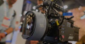 Close-up of a professional ARRI film camera with a large lens labeled "16," surrounded by blurred people and bright lights in the background, suggesting an event or exhibition setting.