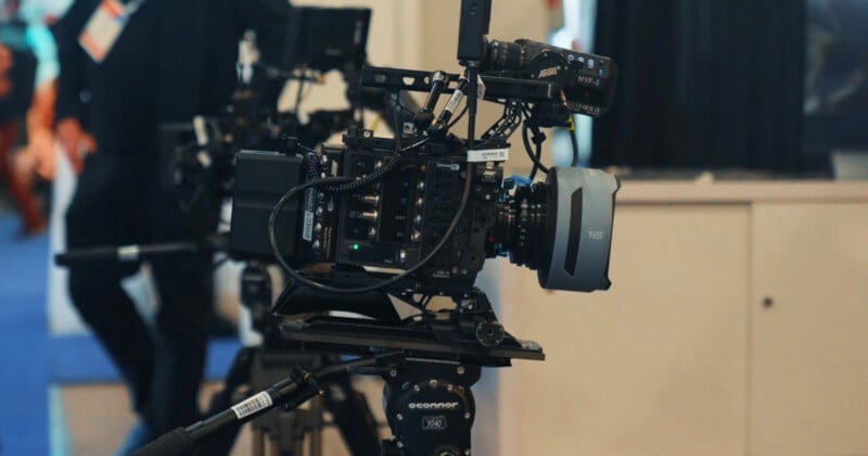 A professional video camera mounted on a tripod is set up indoors, with another camera blurred in the background. Various cables and microphones are attached to the main camera, ready for filming.