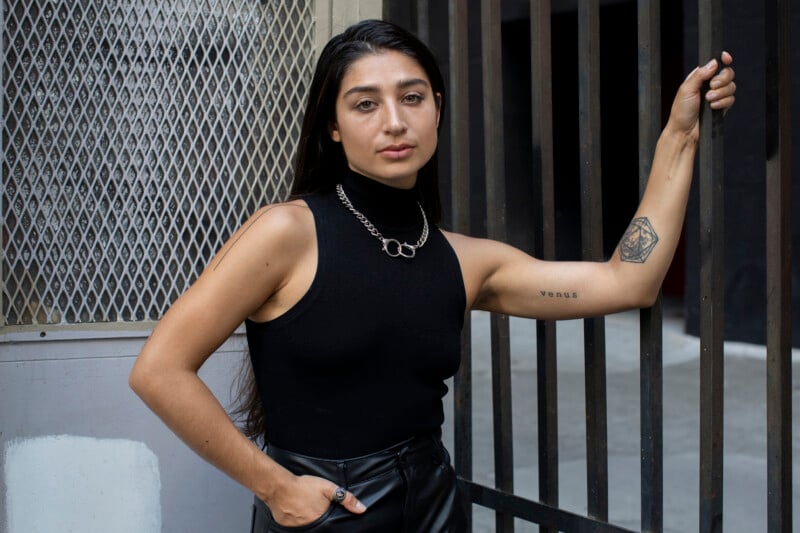 A woman with long dark hair wearing a black sleeveless turtleneck and chain necklace stands by a metal gate, with one hand in her pocket and the other resting on the bars. She has a tattoo on her forearm.