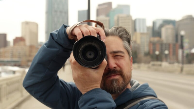 A man with gray hair and a beard is standing on a city street, holding a camera up to his face and taking a photo, with tall buildings blurred in the background.