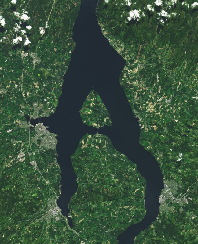 A satellite image shows a large, dark blue lake with irregular shores surrounded by green farmland and patches of forest. Small towns and roads are visible near the lake’s edges.