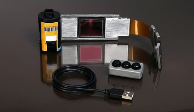 A yellow film canister, a USB cable, a small rectangular device with three black buttons, and a metallic device with a ribbon cable are arranged on a reflective dark surface.