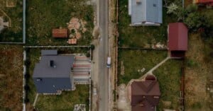 Aerial view of a narrow road with a parked white van, flanked by houses with yards, green grass, and scattered items, separated by fences.