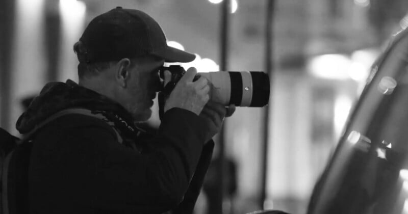A person wearing a cap and a backpack is taking a photo with a large camera lens in a city at night; the image is in black and white.