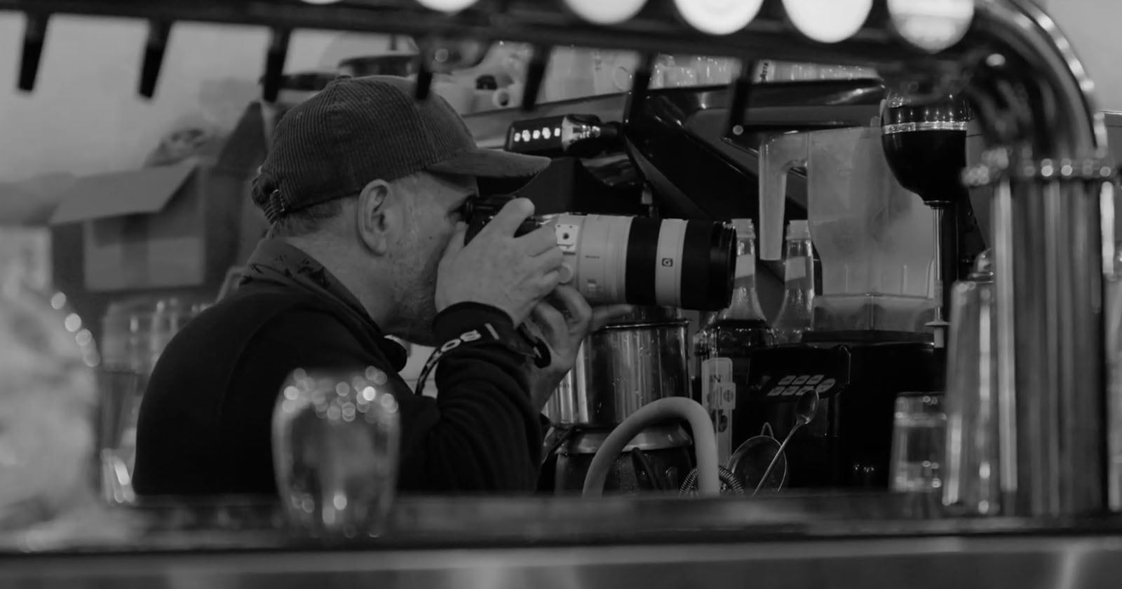 A person wearing a cap and dark clothing holds a camera with a large lens, aiming it while seated at a counter surrounded by coffee shop equipment and glassware. The image is in black and white.