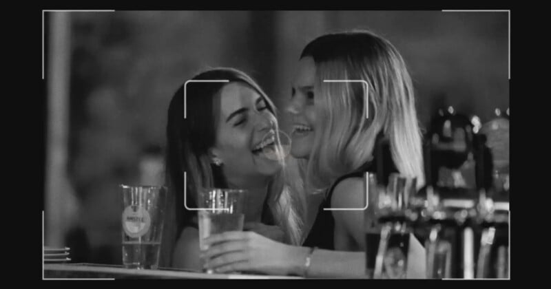 Black and white photo of two women at a bar, smiling and laughing while holding drinks. A camera viewfinder overlay frames their faces, and glasses are visible on the counter in front of them.