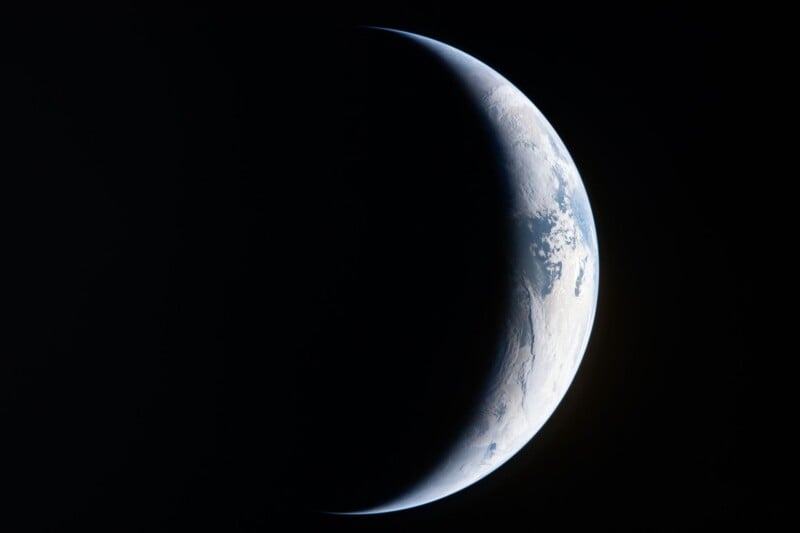 A crescent-shaped view of Earth from space, with sunlight illuminating the right edge and most of the planet in shadow, highlighting the curvature and atmospheric haze.