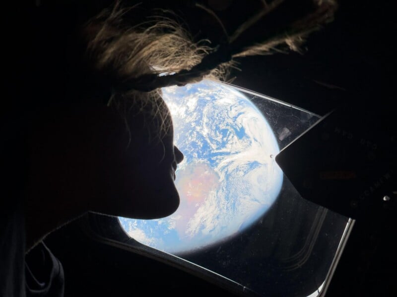 A person with hair floating in microgravity looks out a window at Earth from space, with the planet's blue and white surface visible through the glass.