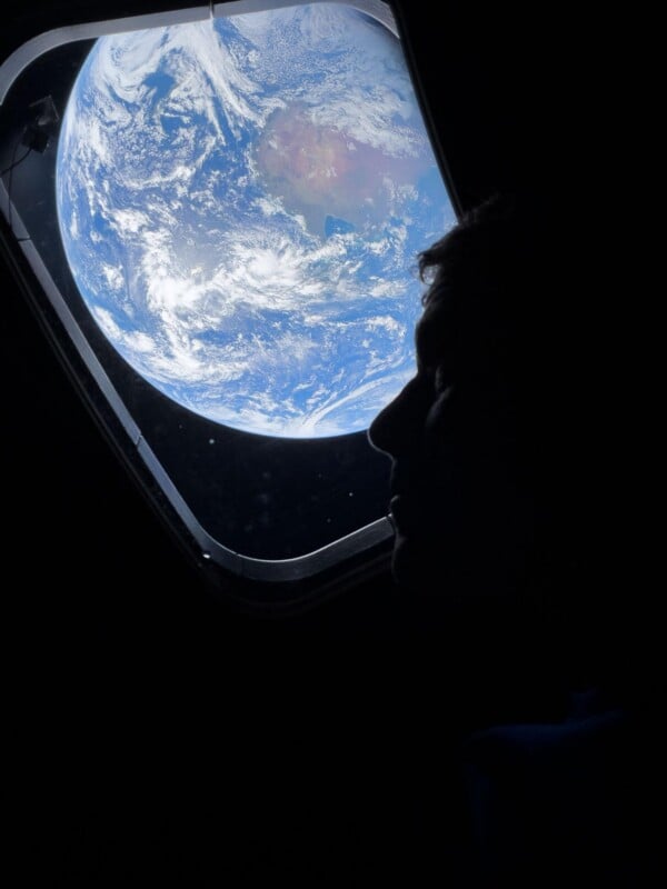 A person’s silhouette looks out a spacecraft window at Earth, which appears blue and white with clouds and continents visible against the blackness of space.
