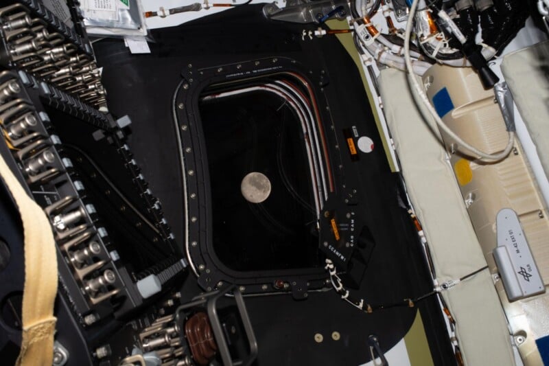 A view of the Moon seen through a window of a spacecraft, surrounded by various cables, controls, and machinery inside the spacecraft.