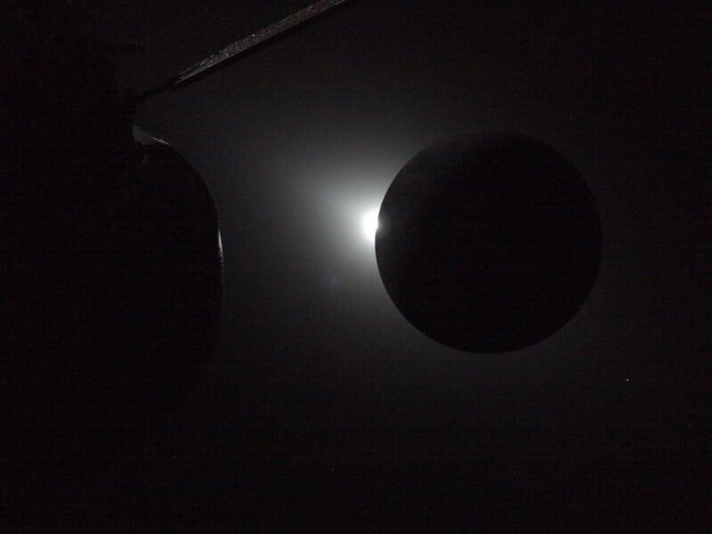 A dark circular object is silhouetted against a bright light source in space, possibly the sun, partially obscured by the object. The image has a dramatic contrast with mostly black and some soft white glow.