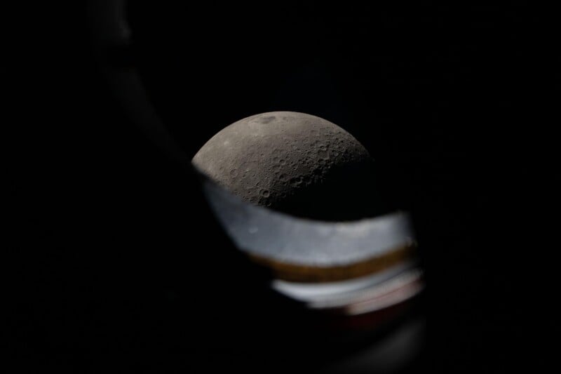A close-up view of the moon’s cratered surface framed by a dark, blurry foreground, possibly taken through a spacecraft window against the blackness of space.