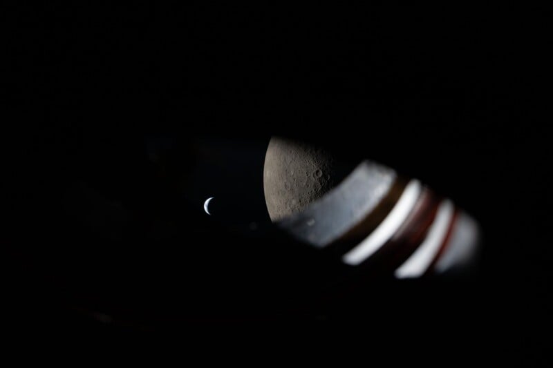 A crescent-shaped Earth is visible in the dark sky above the Moon’s surface, as seen through a spacecraft window with parts of the window frame in the foreground.