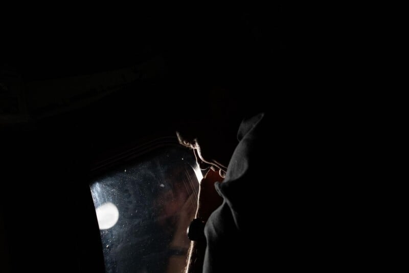 A silhouette of a person in low light, facing a small window with reflections, partially illuminated by light coming from outside the window. The surroundings are mostly dark.