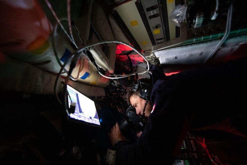 An astronaut wearing a headset operates controls and looks at a screen inside a dimly lit, equipment-filled spacecraft. Cables and electronic panels surround the person, illuminated by red and white lights.