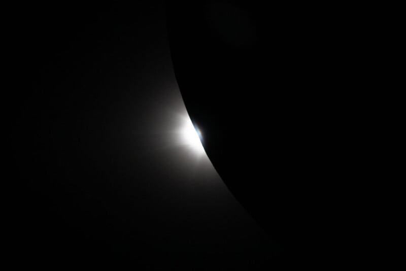 Un brillante rayo de luz se asoma detrás de una gran curva negra, que representa un eclipse solar, con el borde de la corona de la luna claramente visible contra un cielo negro.